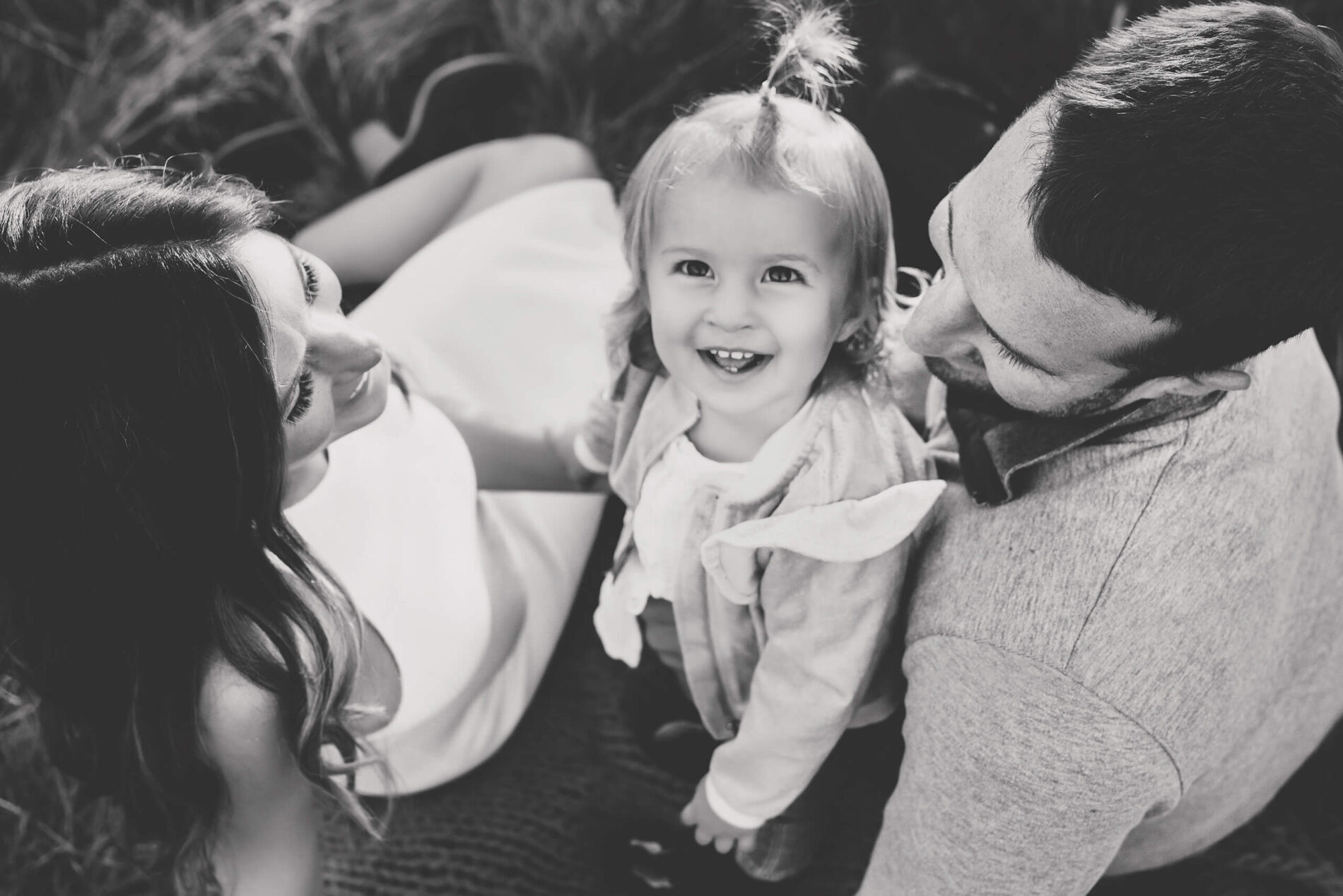 A black and white portrait of a family. Focus is on the smiling young girl looking straight into the camera standing between her parents