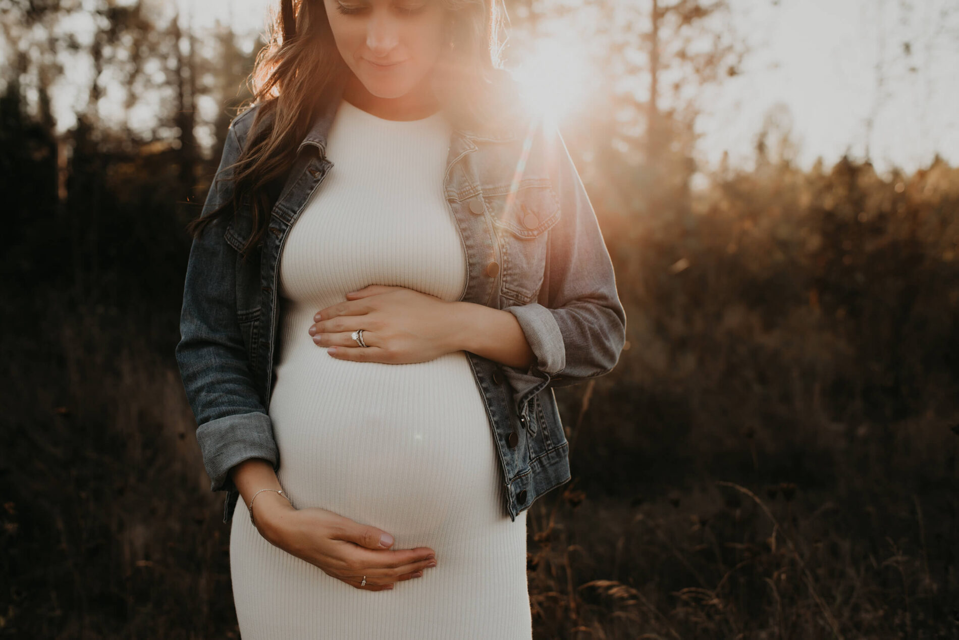 A pregnant woman during photoshoot in Redmond, WA, in a white dress in a meadow with sun rays shining through the trees in the background