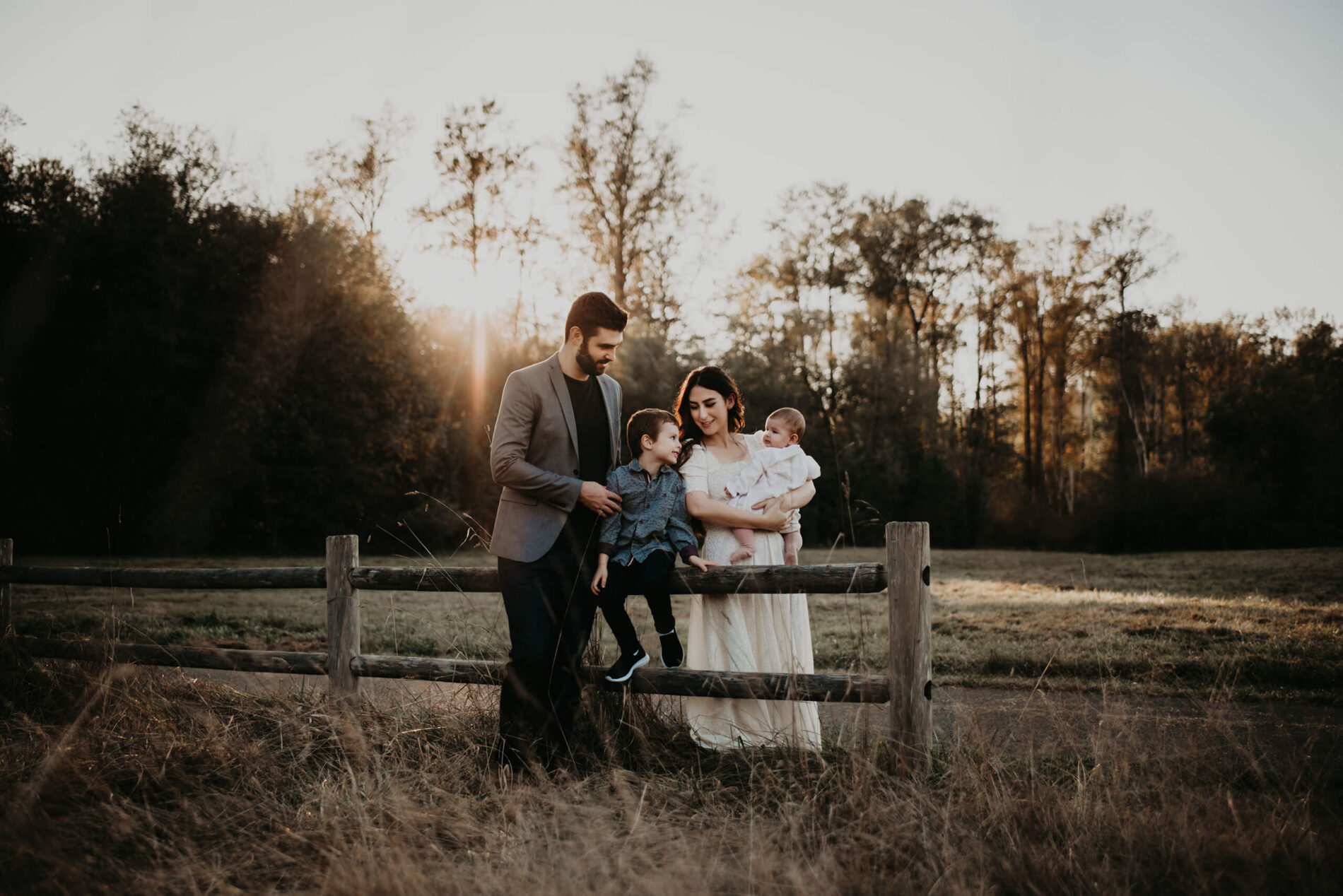 A family portrait in a beautiful field during sunset, mom holding a toddler daughter with her son sitting on a fence, hugged by her husband