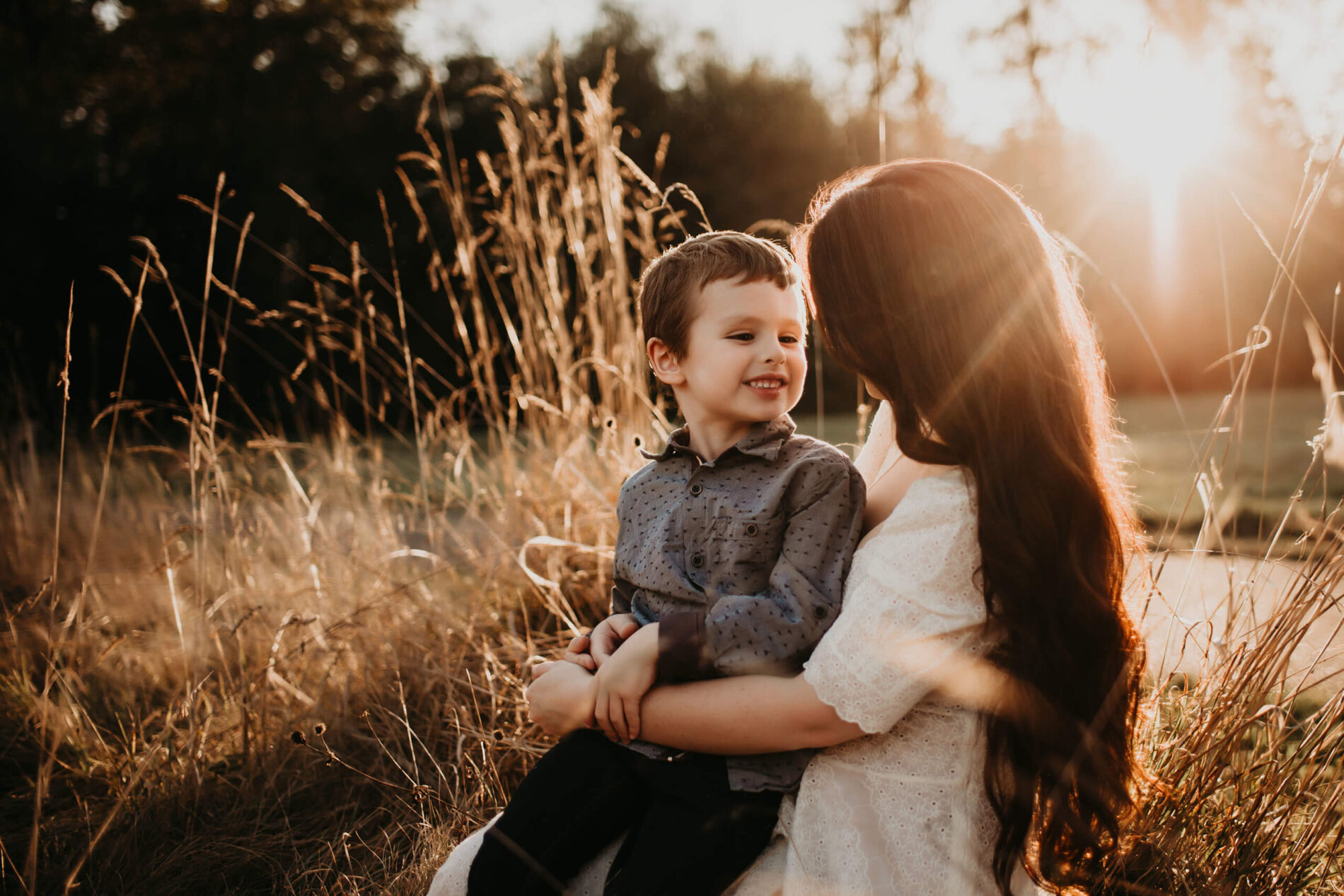 A young boy, sitting in his mom's lap in a field with tall grass, during beautiful golden light of the sunset