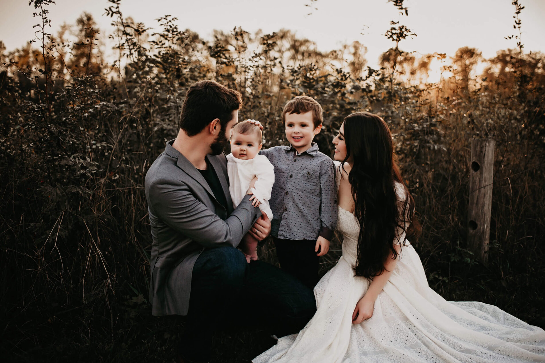Family posing in a field of tall grass, dad holding a toddler daughter, with his son and wife next to him