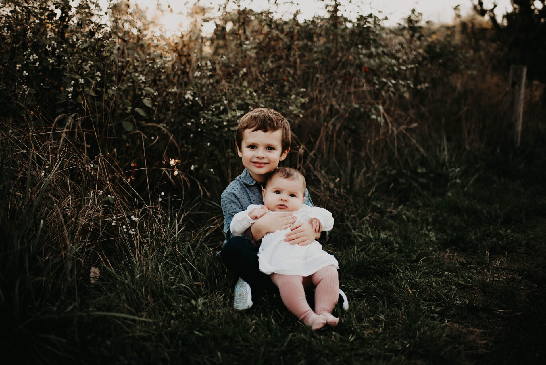 A young boy sitting in a field and holding his toddler sister in his arms