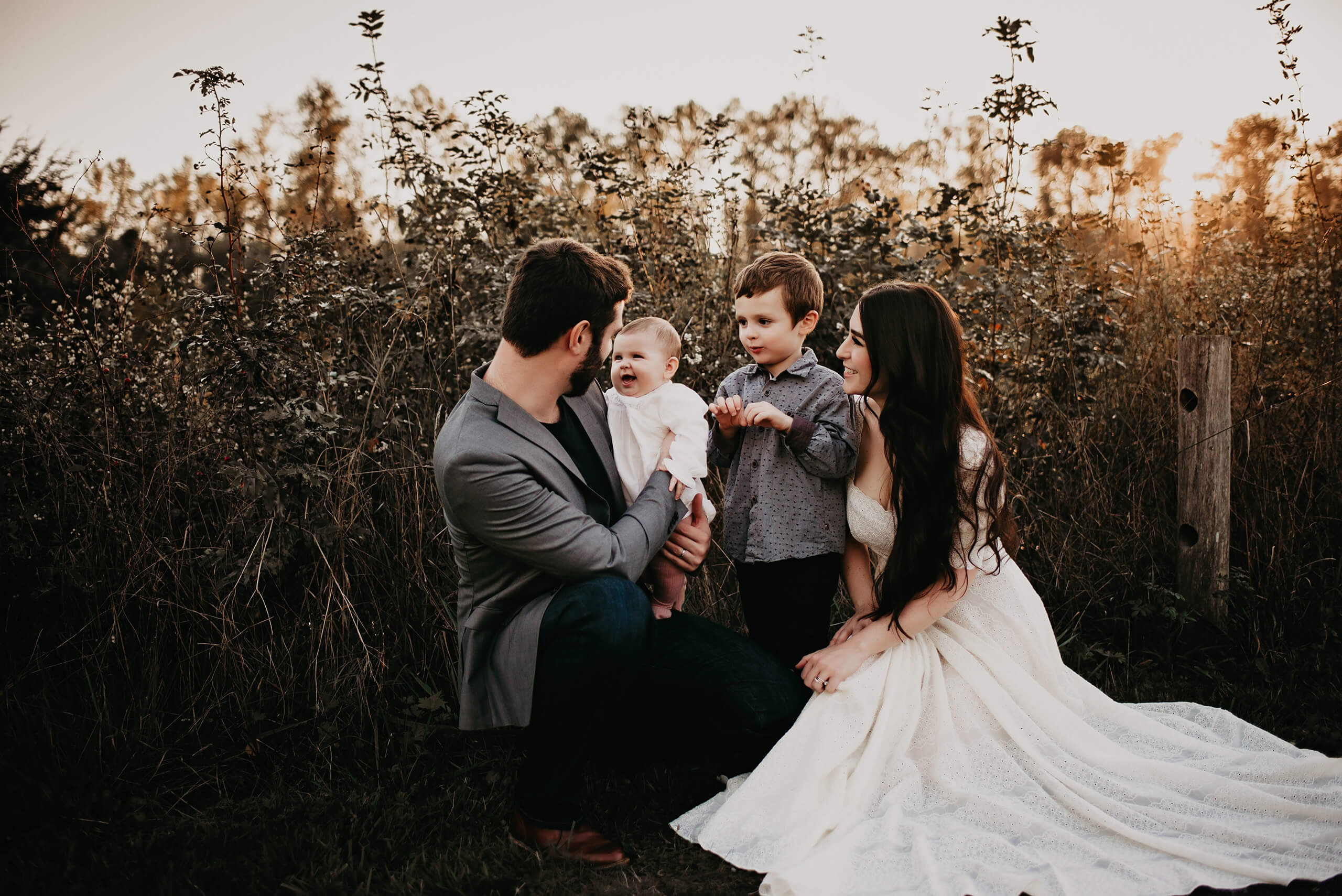 A man holding his toddler daughter who is smiling, with his wife and son standing next to him in a field of tall grass