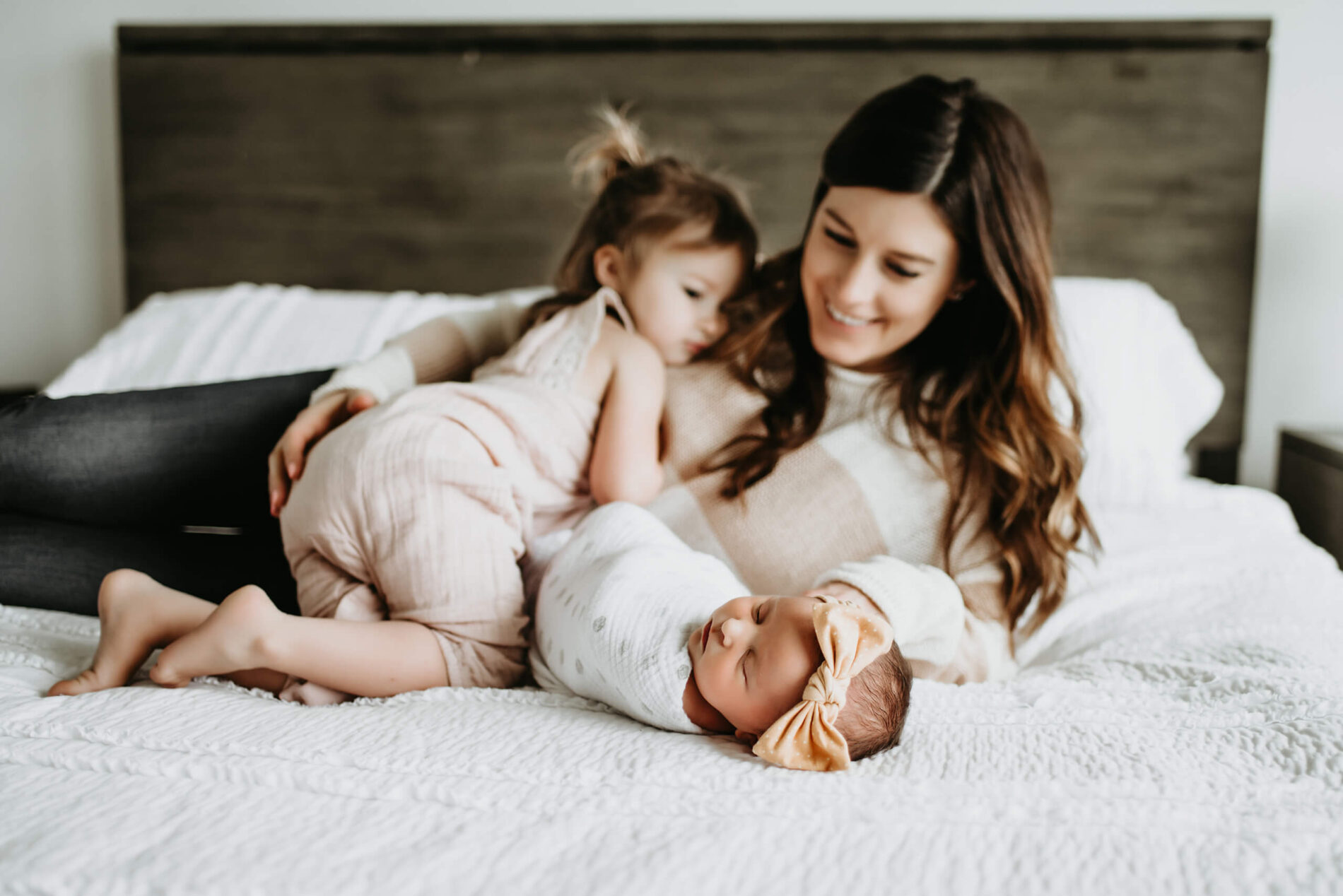 Mom playing with her toddler daughter and newborn daughter on a large bed