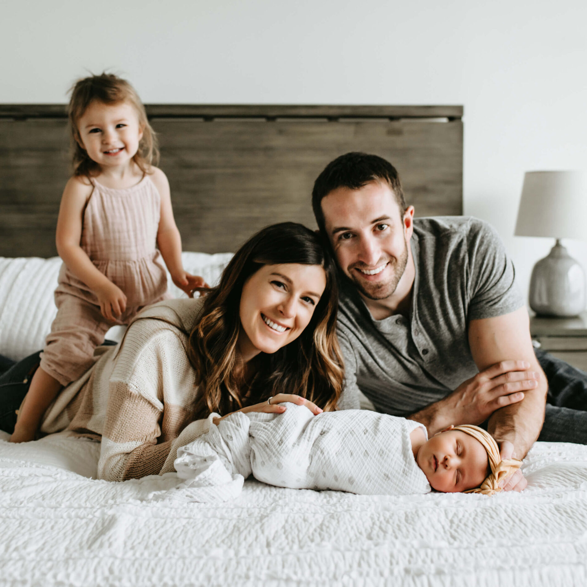 Mom and dad lying on the bed smiling, with sleeping newborn daughter in front of them, and older daughter sitting on top of mom
