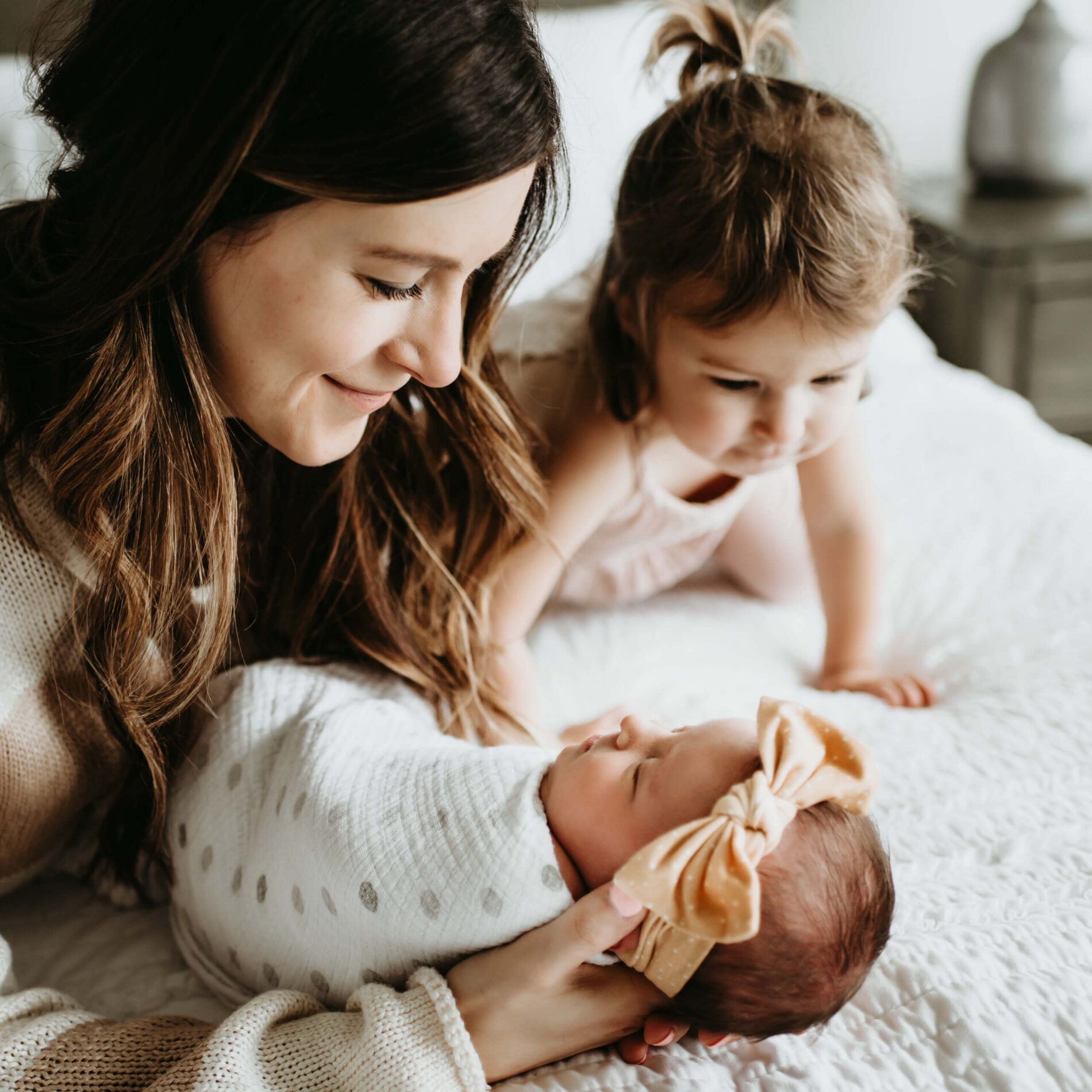Mom lying on a bed, holding hew newborn daughter in her hands, while toddler daughter is crawling on the bed