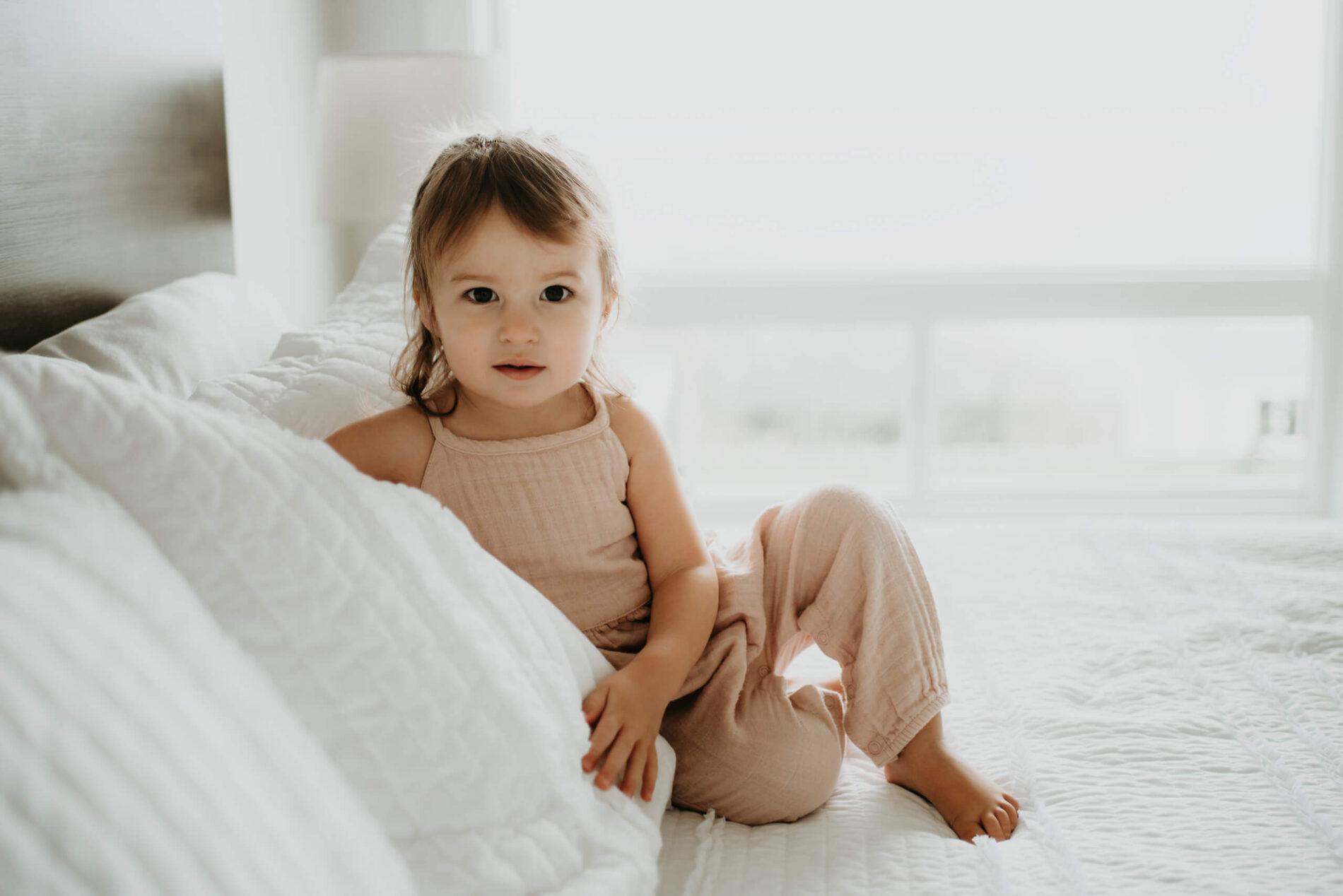 A girl leaning on pillows on a large bed, with light from large windows flooding the room