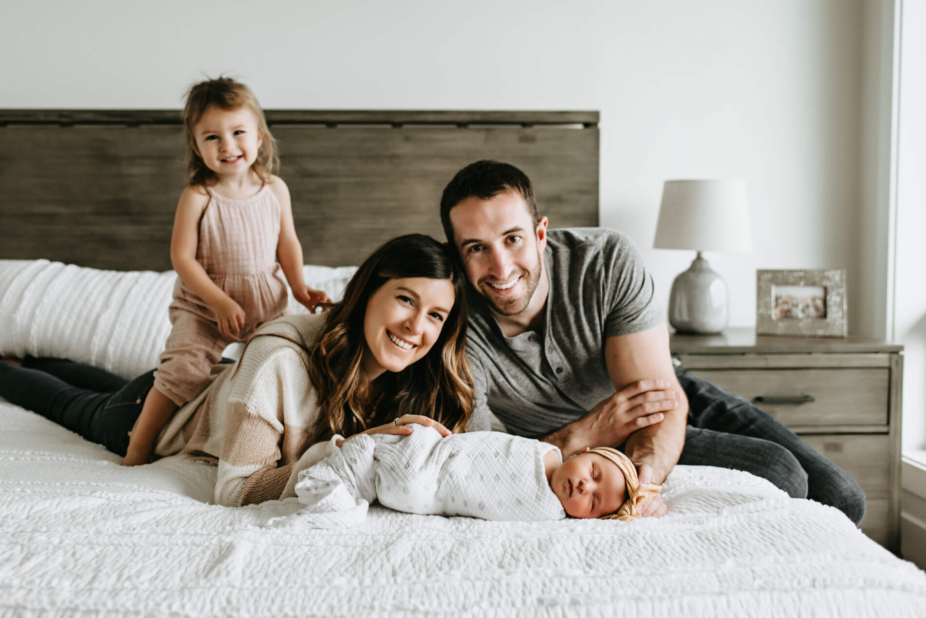 Parents lying on the bed of their bedroom during newborn photoshoot with infant daughter and toddler daughter sitting on mom