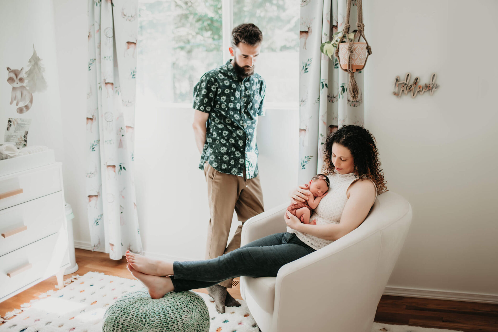 Mom holding hew newborn daughter in a rocking chair in a nursery with dad watching