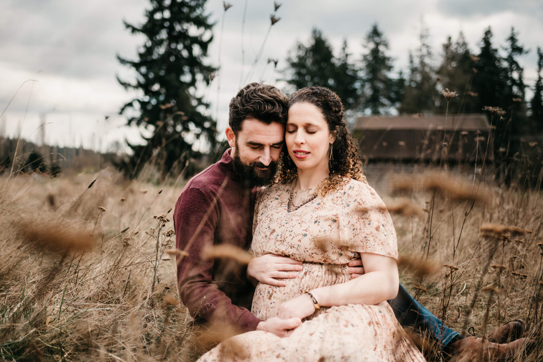 Pregnant woman sitting in a field with eyes closed, hugged by her husband from behind