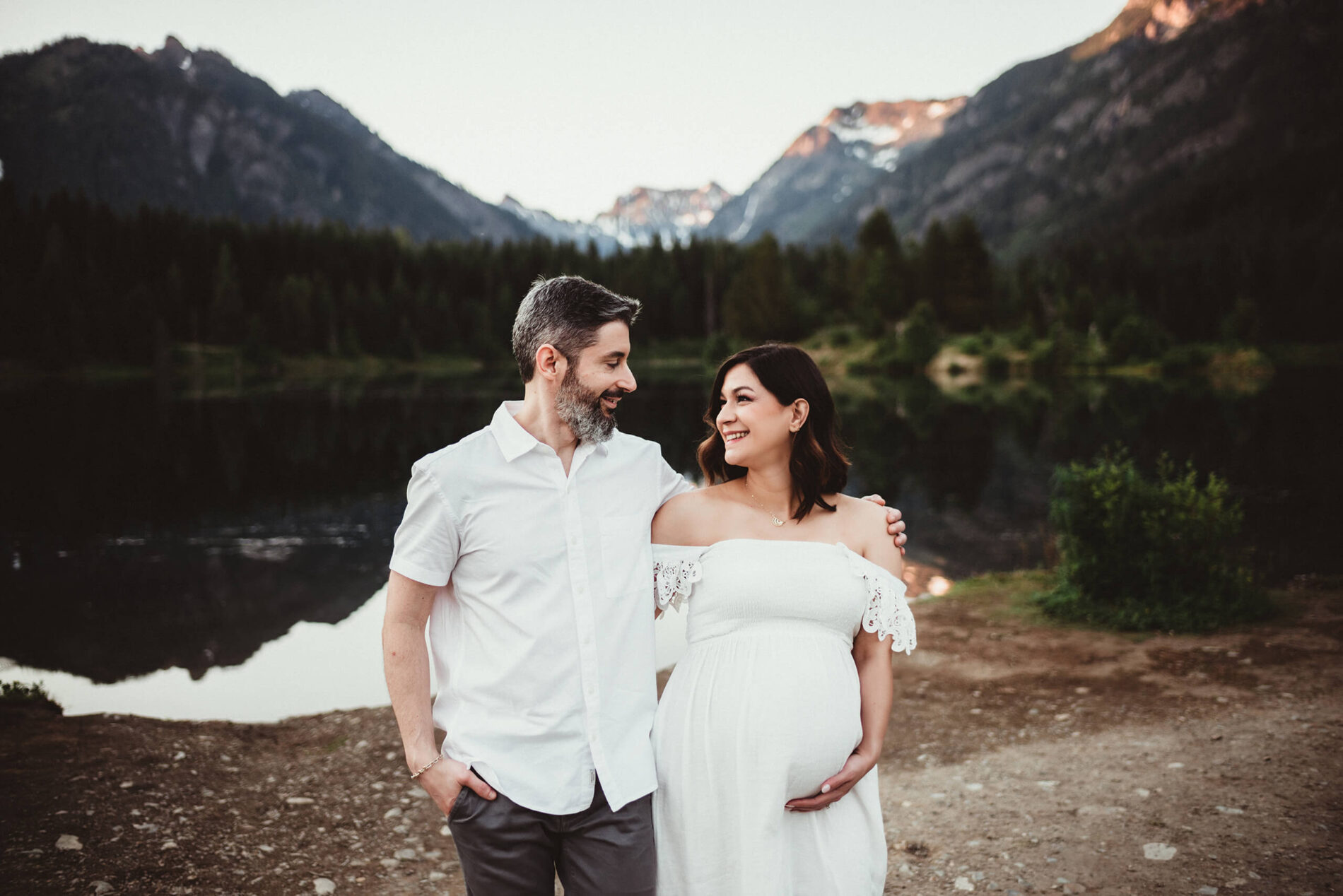Maternity photoshoot at Snoqualmie Pass, pregnant woman in a white dress smiling and hugging her husband, stunning mountains in the background