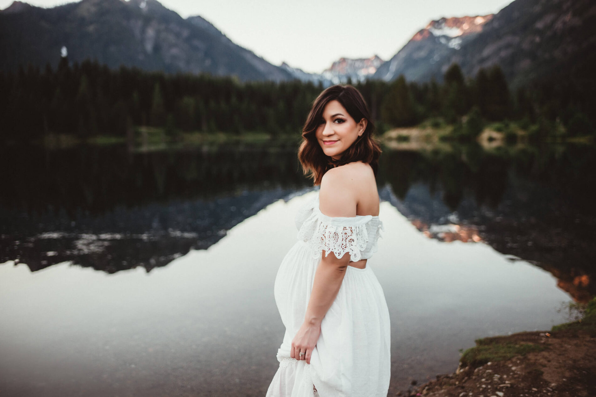 Pregnant woman in a white dress standing next to a lake with a beautiful mountain backdrop
