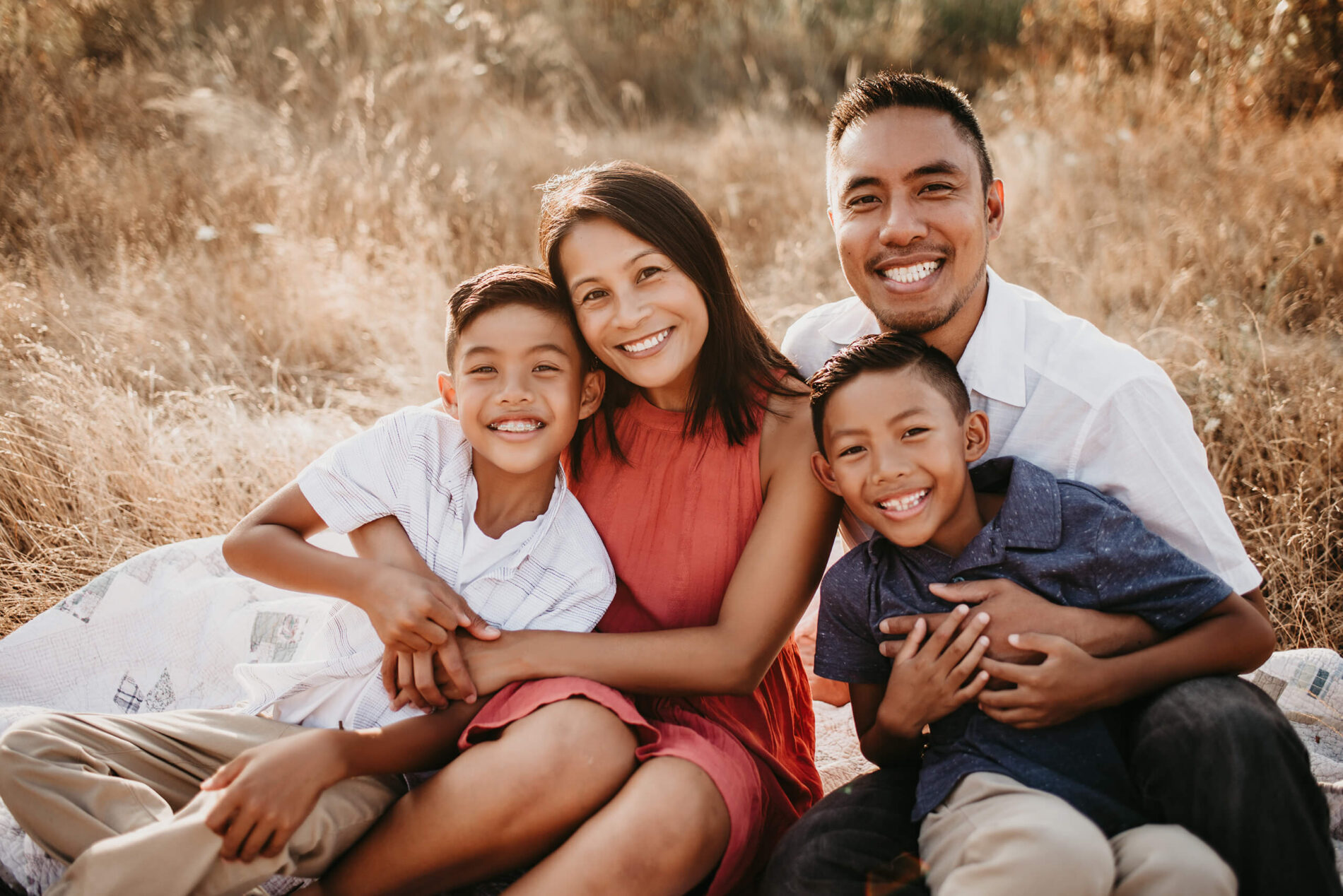 Mom, dad, and two sons sitting in a field hugging and smiling