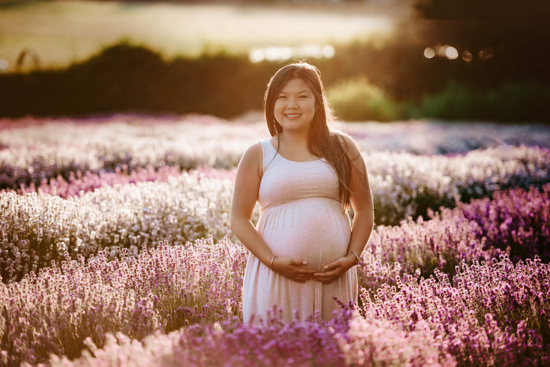 Pregnant woman in a white dress standing in the middle of a stunning lavender field and smiling