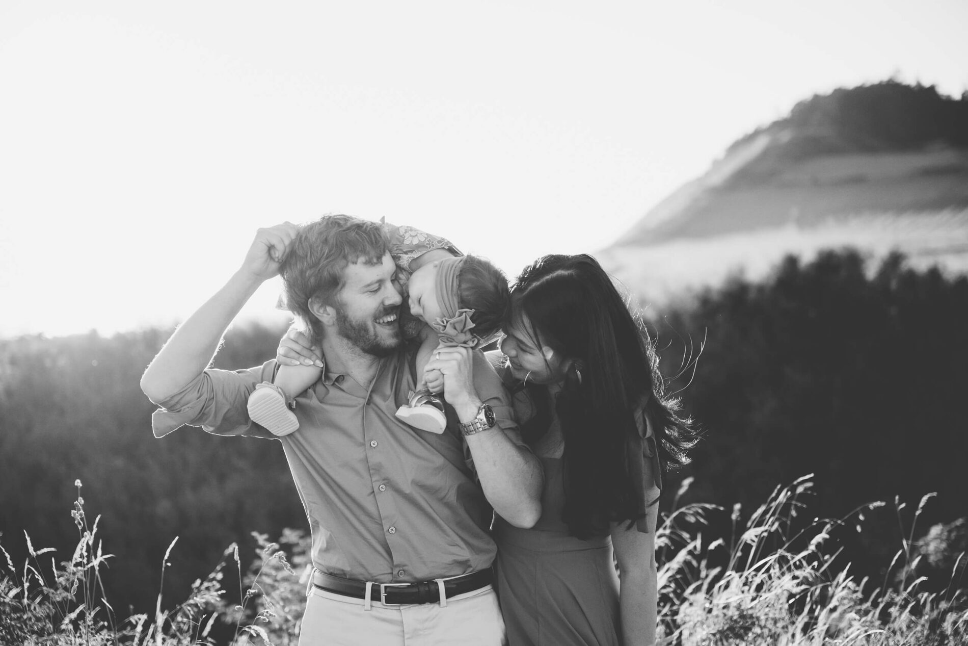 Dad holding his baby girl on his shoulders, while mom is walking besides them in a field