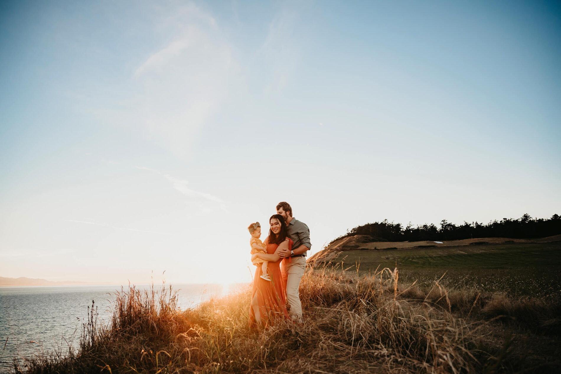 Mom holding her toddler daughter in her hands, being hugged by her husband during sunset and overlooking an ocean