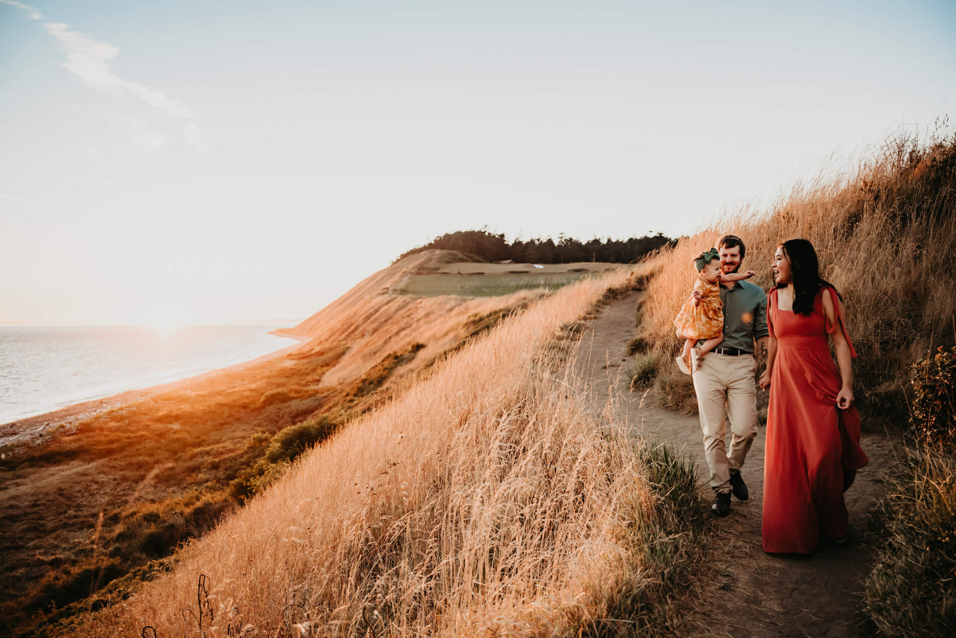 Dad holding his toddler daughter in one hand, and holding his wife's hand in another hand, during sunset