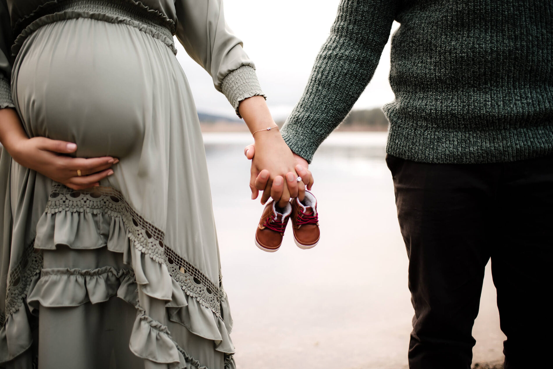 Husband and wife holding hands and tiny baby shoes with serene lake in the background during a winter maternity photo shoot