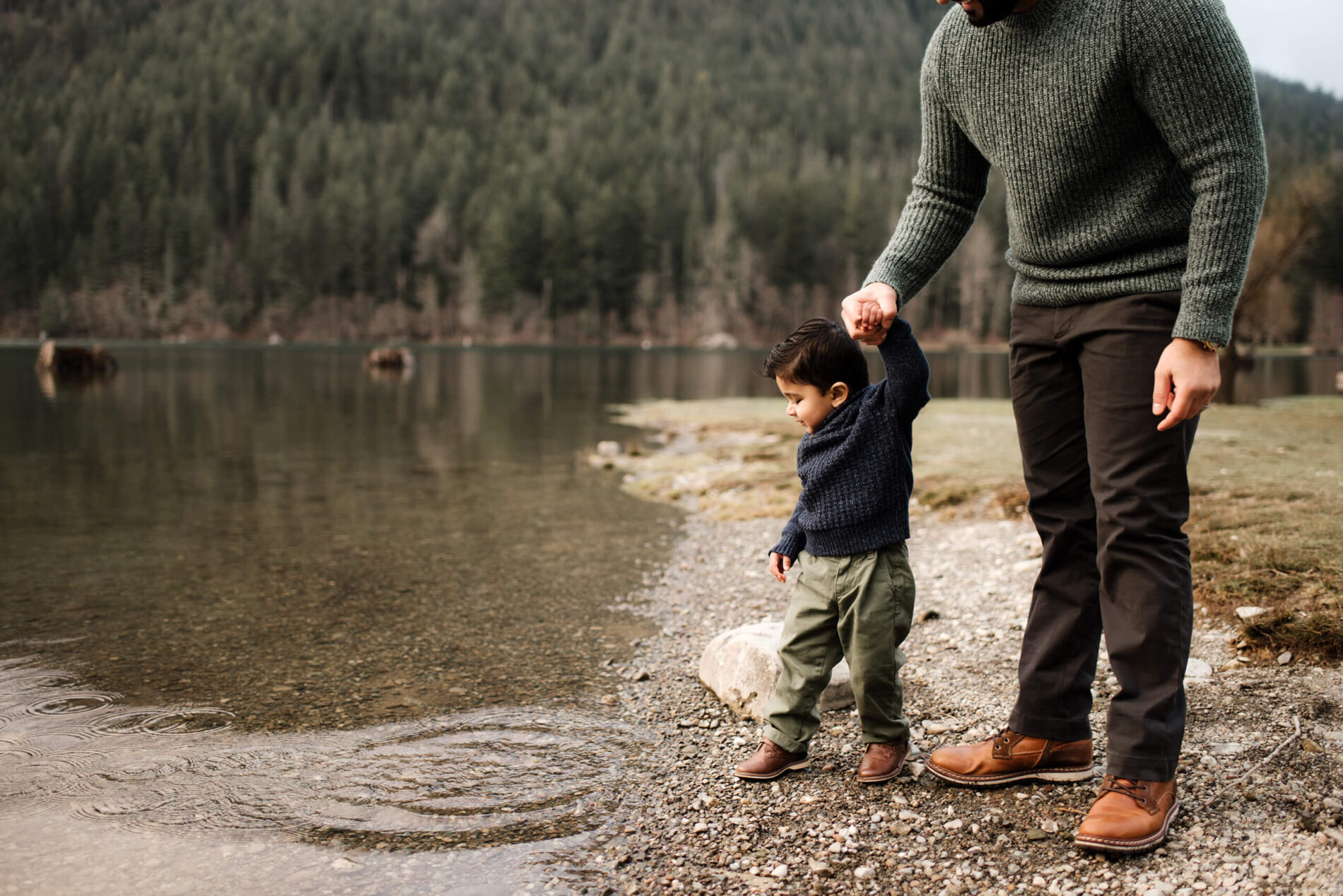Father holding the hand of his son who is throwing rocks into the lake during winter photo shoot