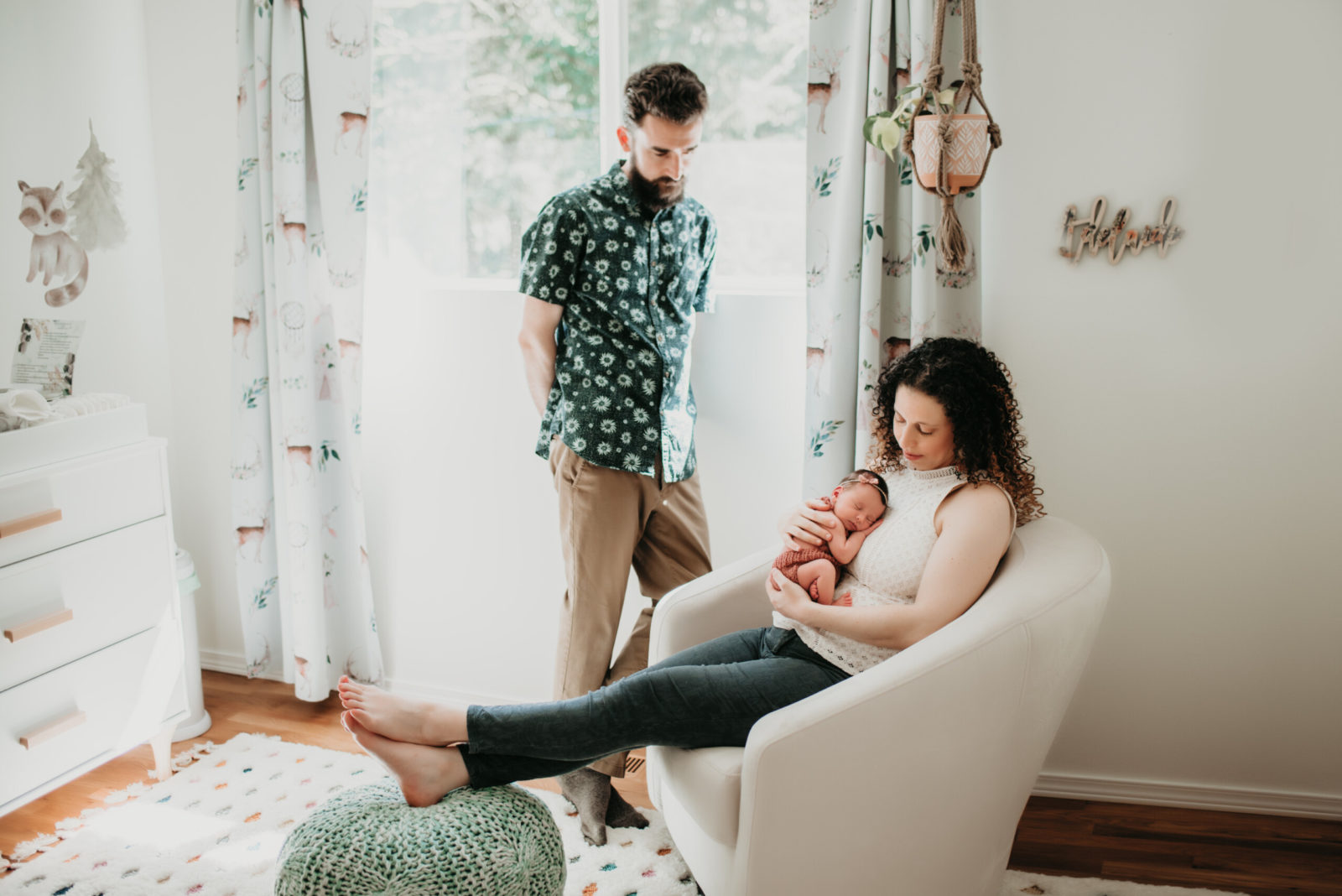 Mom sitting in a nursery rocking chair holding her newborn daughter while dad looks on.