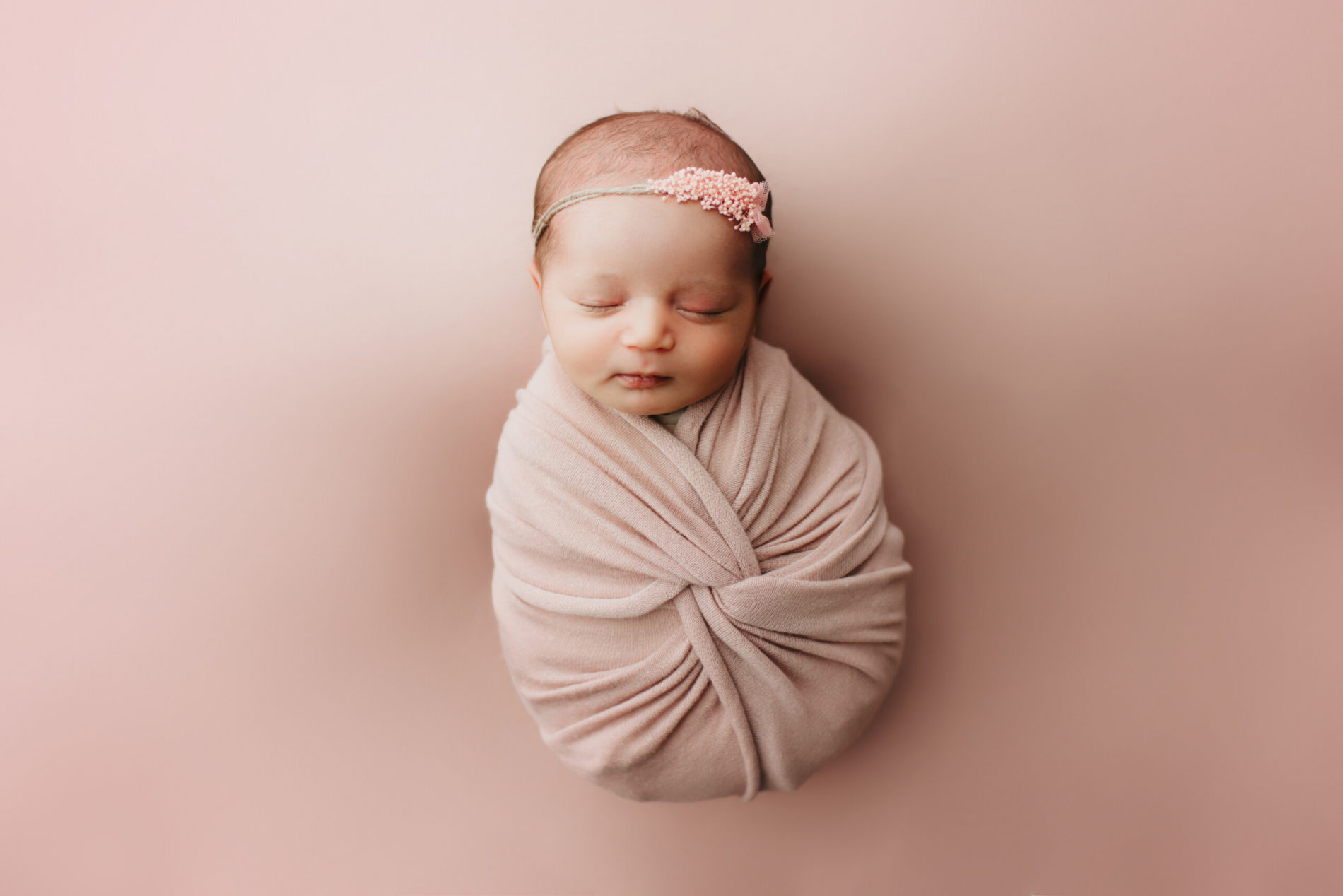 A beautiful newborn girl wrapped in pink wraps, sleeping on a pink background, wearing a floral headband