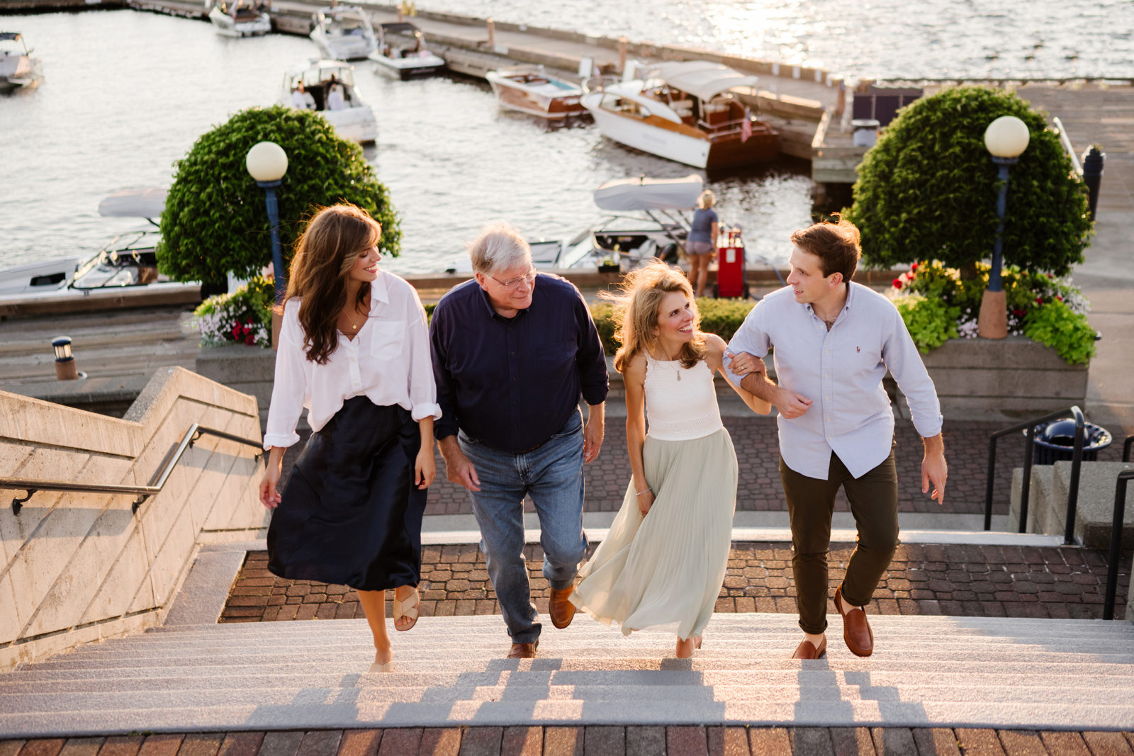 Family photography in Kirkland Carillon Point: father, two adult children, and mom walking up sunlit steps with boats on Lake Washington behind.