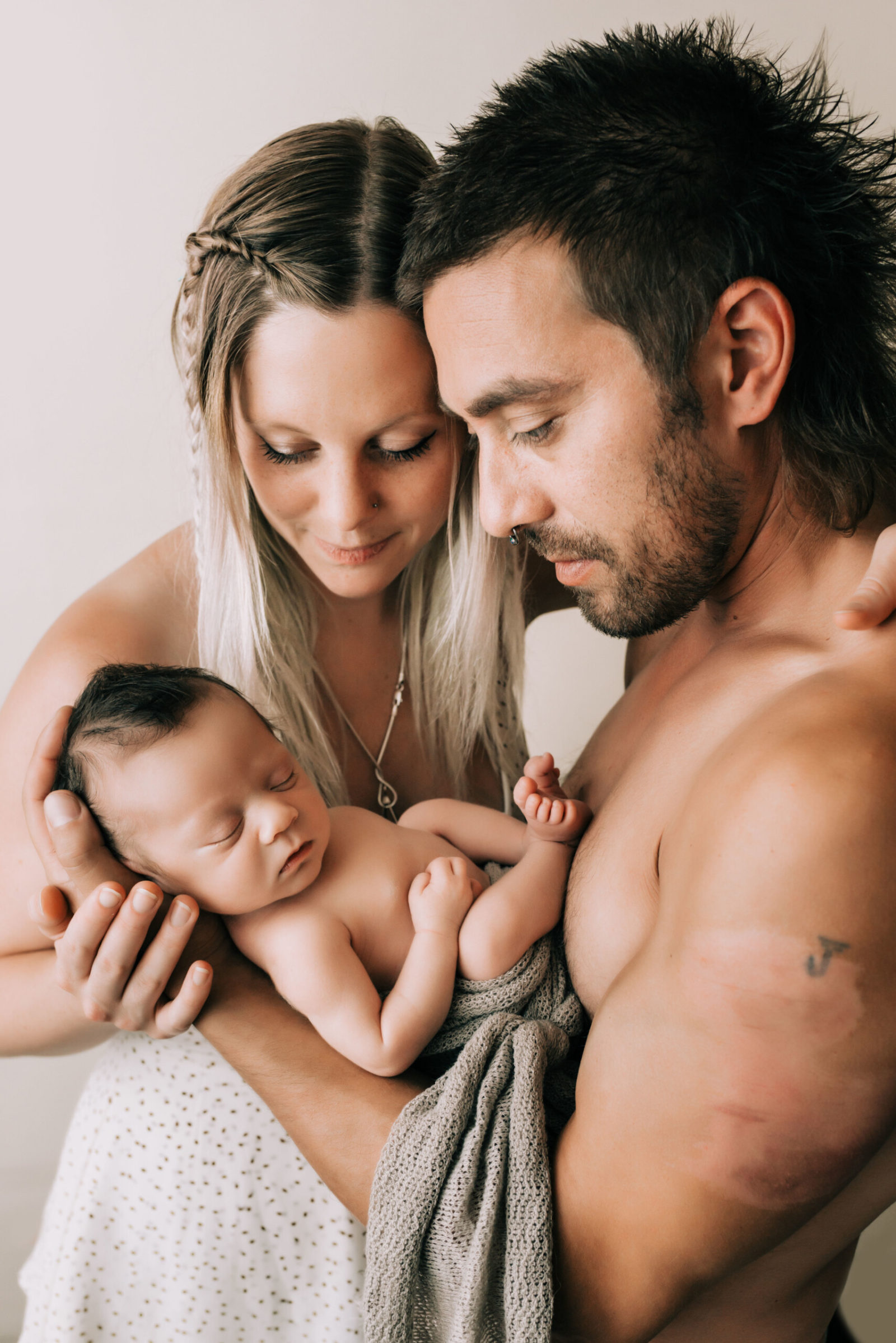 Seattle newborn studio portrait: tattooed dad and braided-hair mom cuddle skin-to-skin, gently cradling their swaddled sleeping baby boy against a soft neutral backdrop