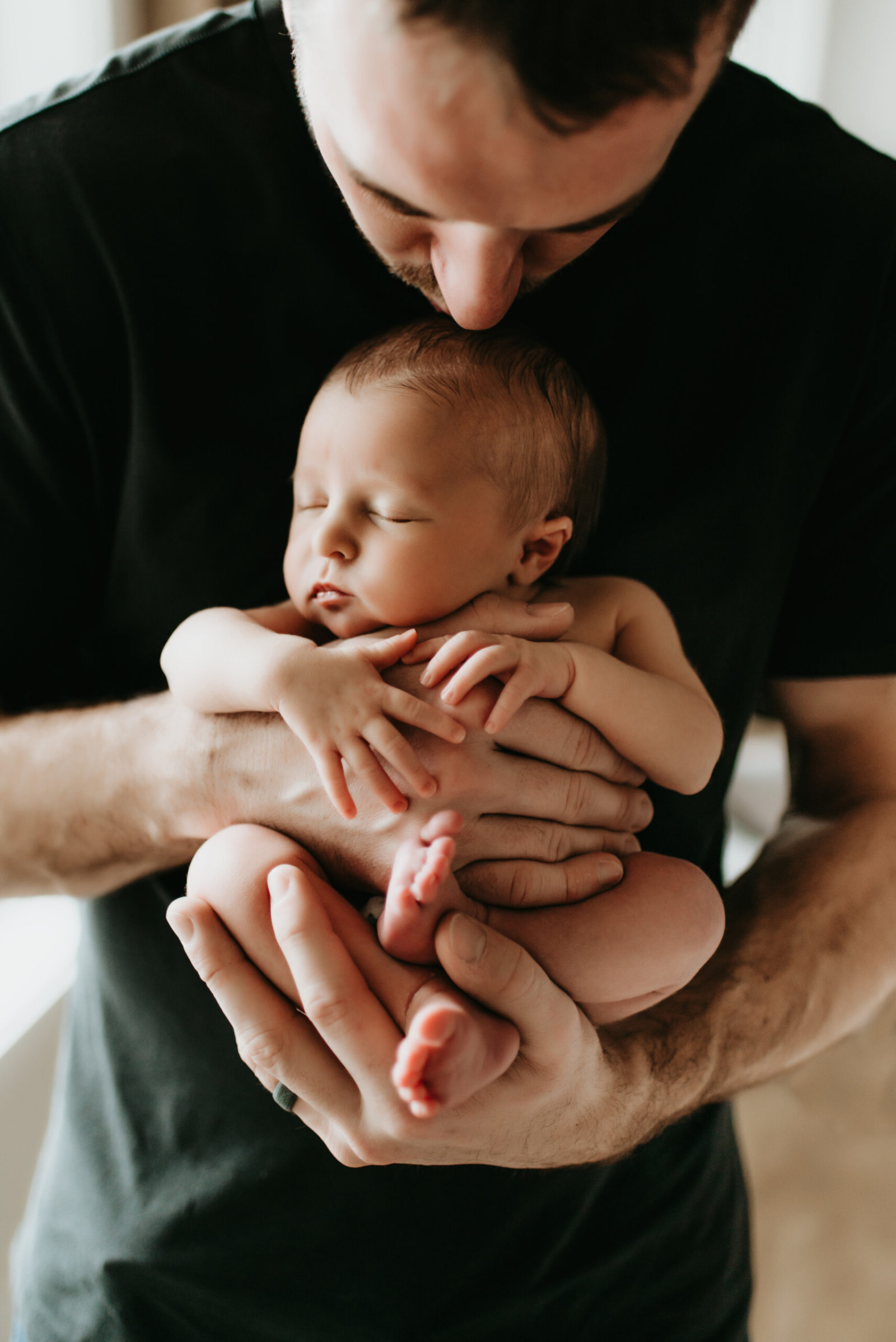Seattle newborn photo: dad in black tee cradles sleeping newborn in both hands, gently kissing baby’s head in soft window light—captured by Lana Sky Photography.