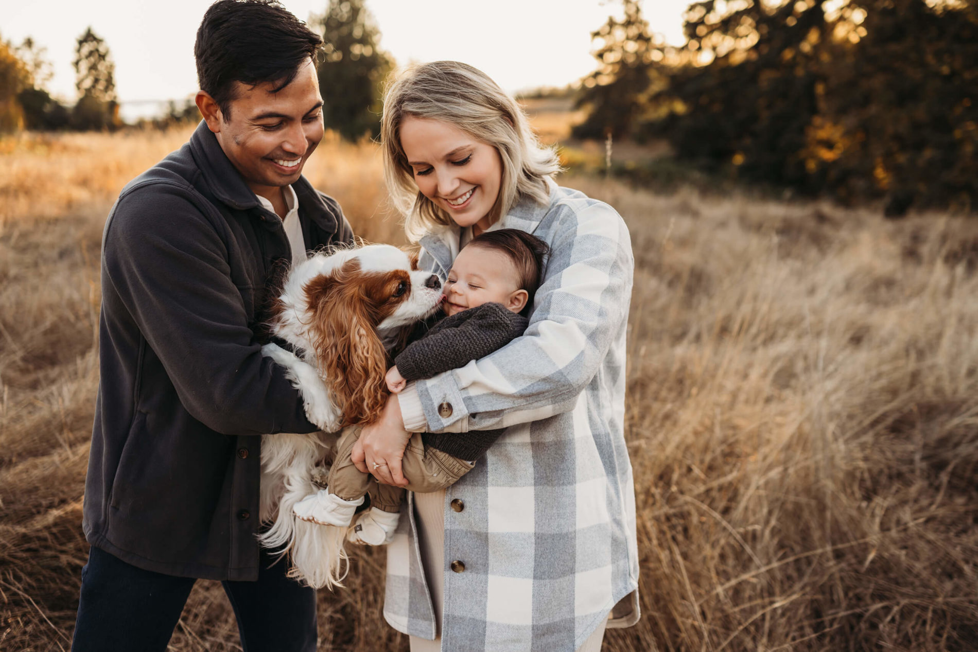 Dad, mom, baby boy and their dog smiling and hugging in a scenic field during sunset in Seattle, WA