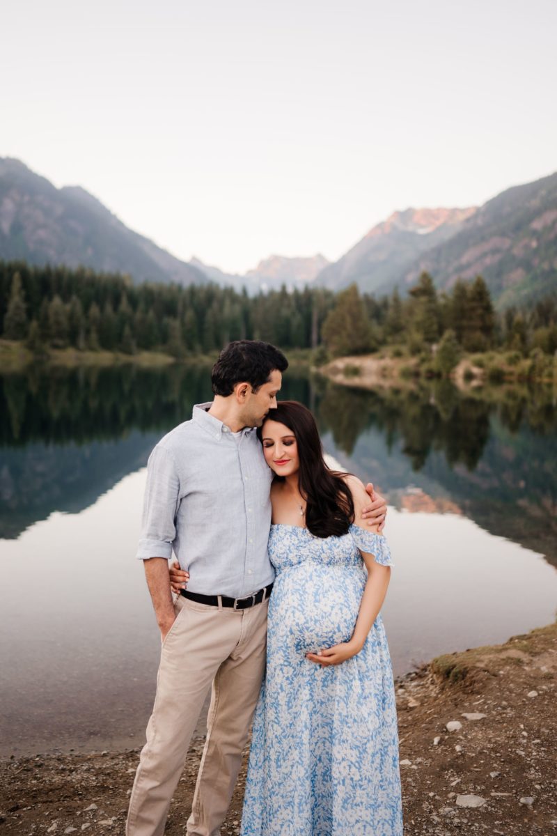 A pregnant woman in a blue dress, being hugged by her husband with lake and mountains in the background
