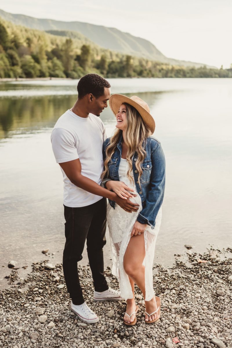 A man hugging his pregnant wife and looking at her lovingly with beautiful lake and mountains in the background