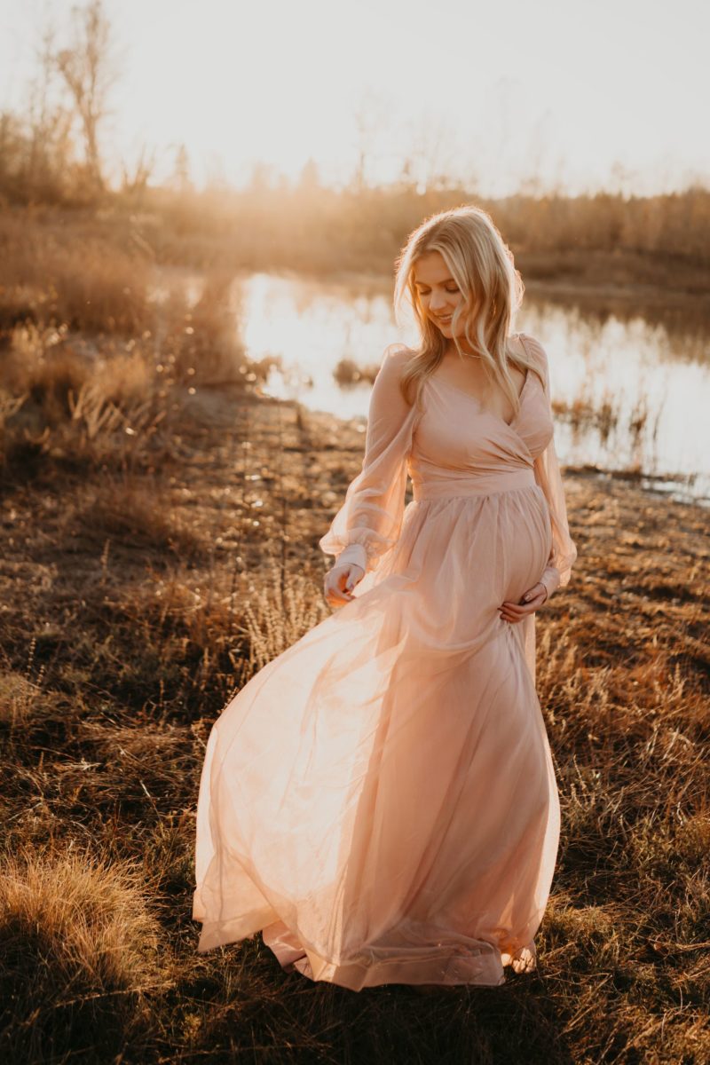 A beautiful pregnant woman in a maternity gown smiling, standing in a field of tall grass during sunset