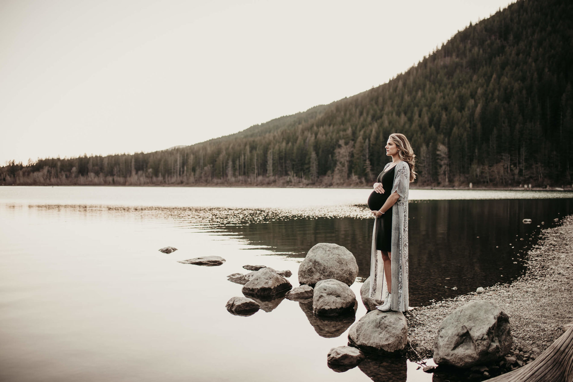 A radiant pregnant woman in a black dress stands gracefully by a tranquil Rattlesnake Lake, framed by majestic mountains in the distance
