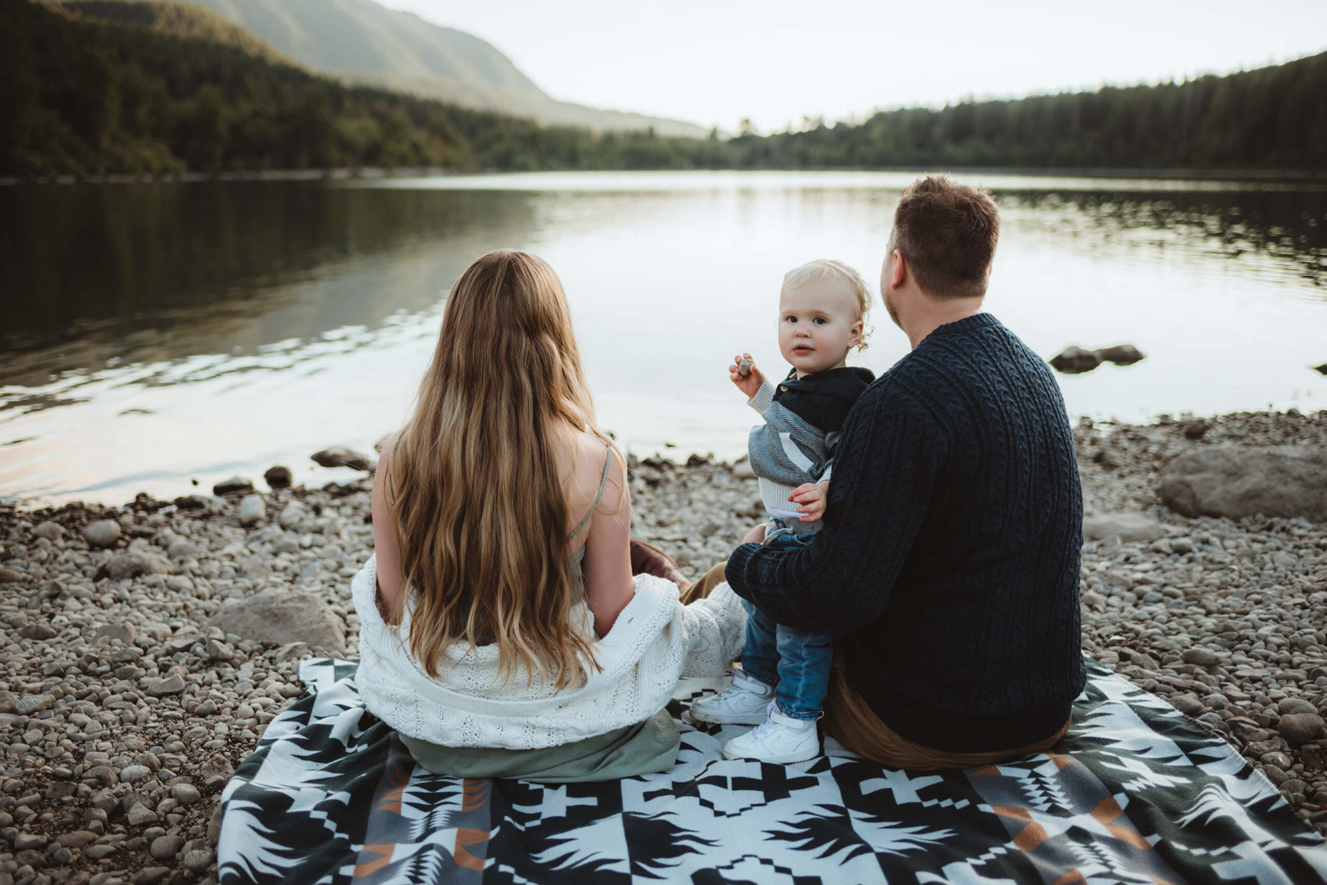In a serene landscape, mom, dad and toddler sitting together looking at a lake