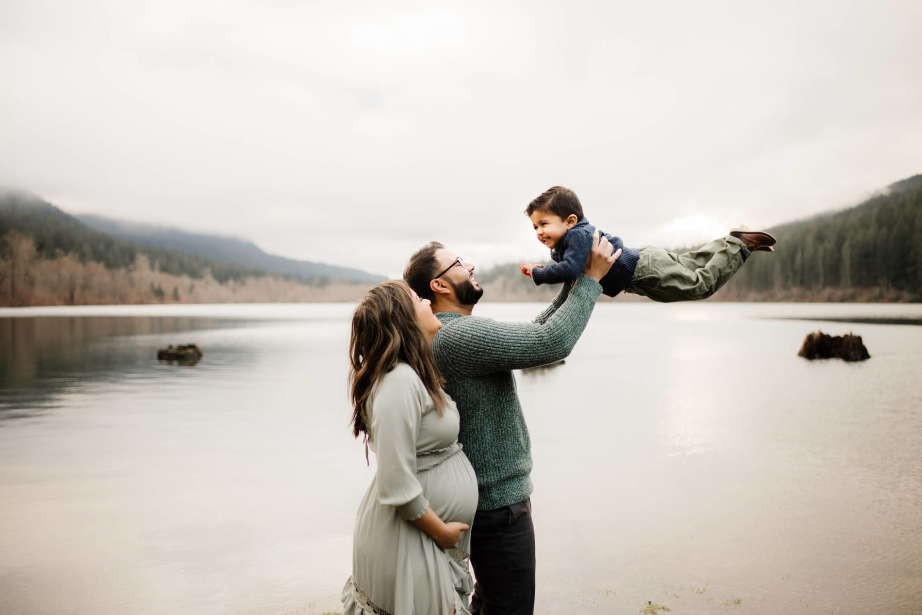 A serene lakeside scene captures the joy and togetherness of a husband, his pregnant wife, and their toddler, standing side by side, with majestic mountains forming a magnificent backdrop.