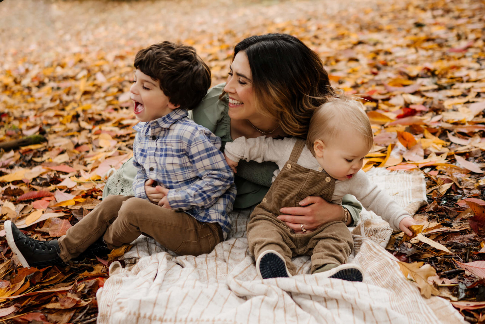 Seattle fall family photography at Washington Park Arboretum: mom in sage jacket hugs laughing preschool son in plaid while baby in corduroy overalls reaches for colorful leaves.