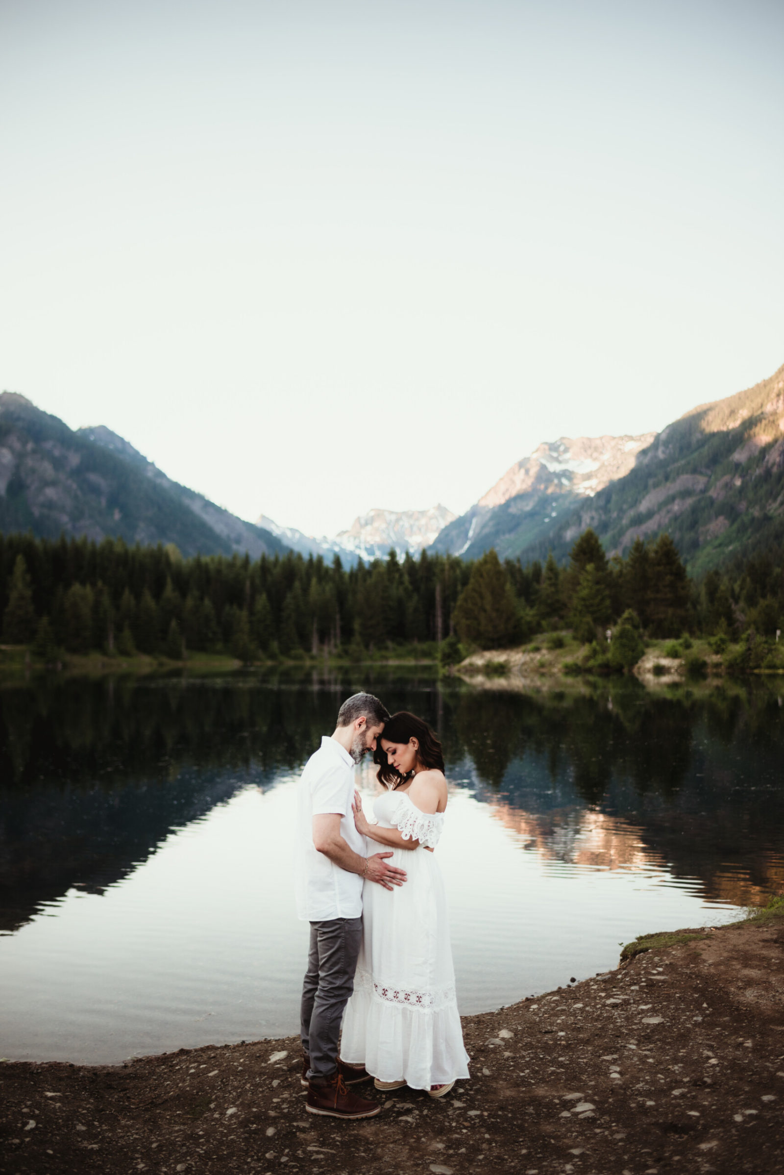 Maternity photoshoot in Seattle area: expectant couple in white attire stands on rocky lake shore, touching baby bump; Cascade peaks and evergreen forest mirror in calm water.