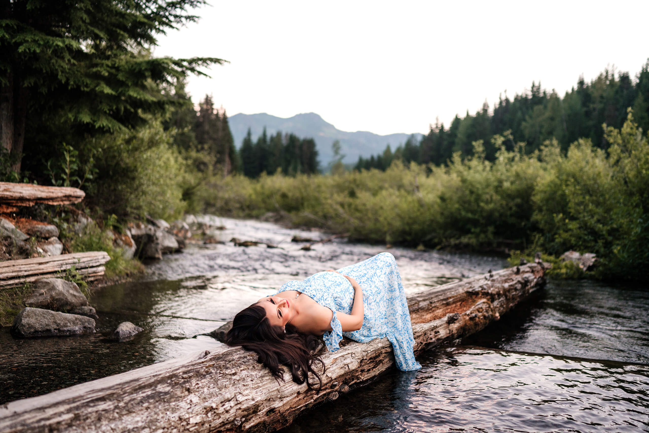Seattle-area outdoor maternity portrait: expectant mom in blue floral gown reclines on a fallen log over an alpine creek with Cascade evergreens and peaks behind—captured by Lana Sky Photography.