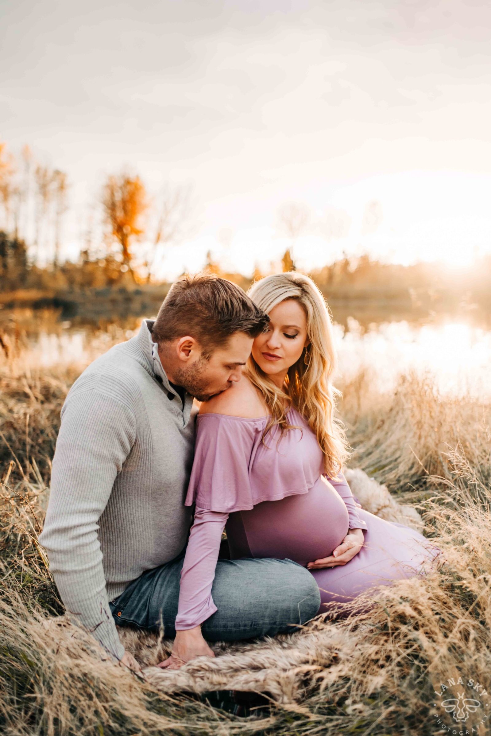 Golden-hour maternity photoshoot in Seattle area: expectant mom in lavender off-shoulder gown sits on a fur throw in a lakeside meadow while partner kisses her shoulder and holds her baby bump.