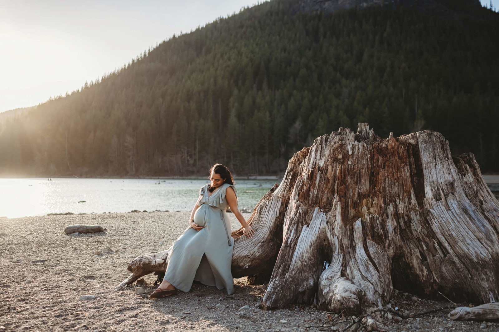 Pregnant woman seated beside a large weathered tree stump with a mountain lake in the background.