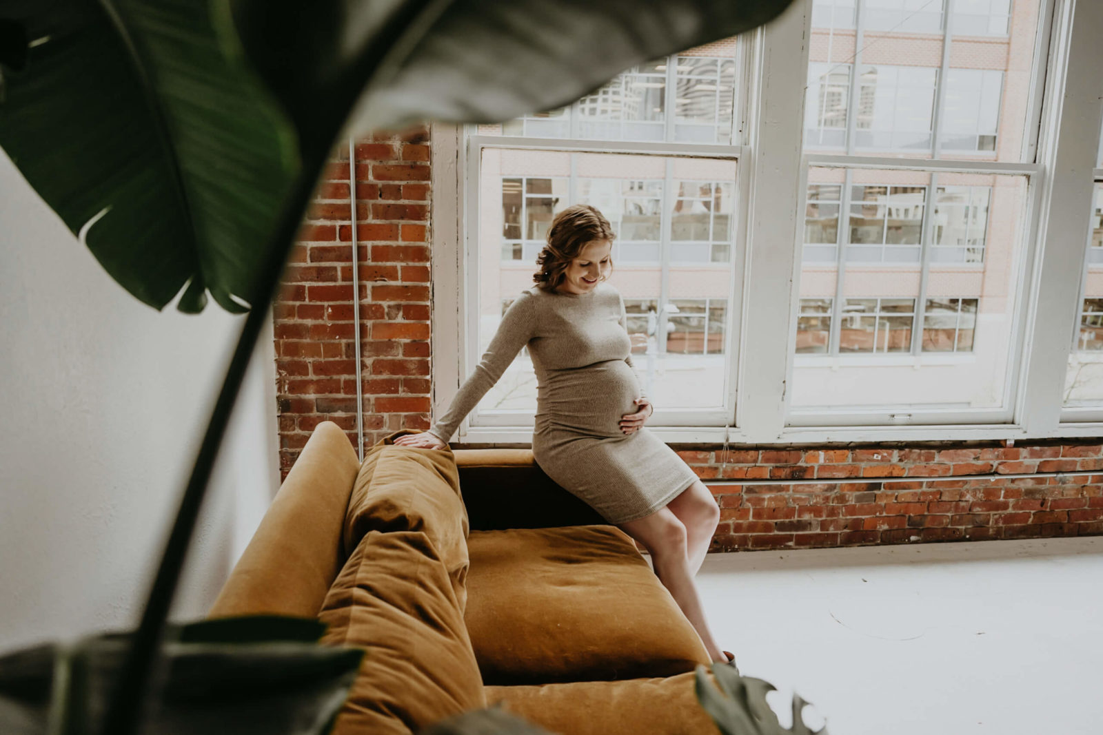 Studio maternity photoshoot in Seattle: expectant mom in taupe knit dress sits on mustard velvet sofa by tall loft windows and brick wall, smiling while cradling her baby bump—Lana Sky Photography.