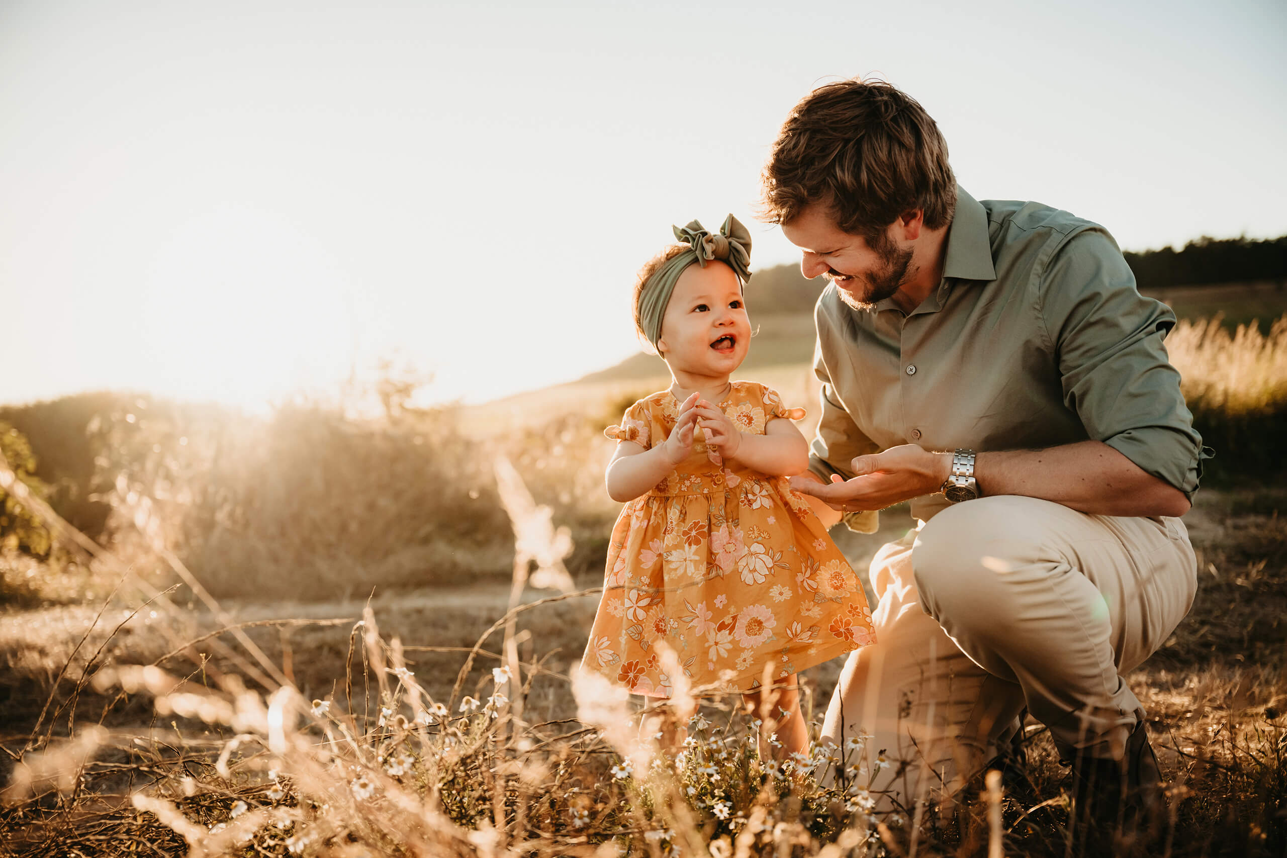 Seattle summer family portrait: dad kneels in golden-hour meadow, laughing with toddler daughter in yellow floral dress and green bow headband—captured by Lana Sky Photography.