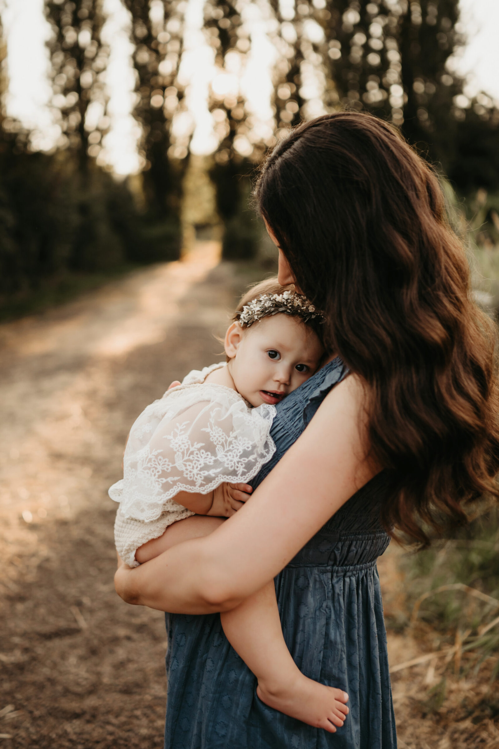 Seattle summer family photoshoot: mother in blue dress cradles and kisses her baby girl, who wears a lace romper and floral headband, on a sun-dappled woodland path.