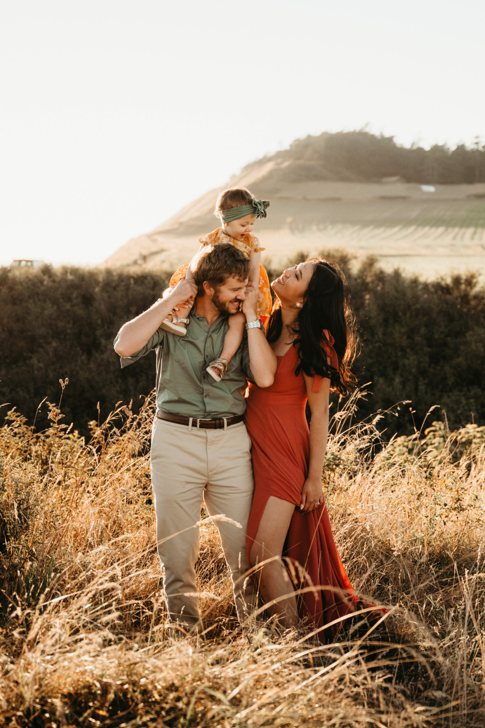 Seattle summer family photoshoot: father carries toddler daughter in yellow floral dress and green bow on his shoulders while mother in red slit dress smiles in a golden meadow at sunset.