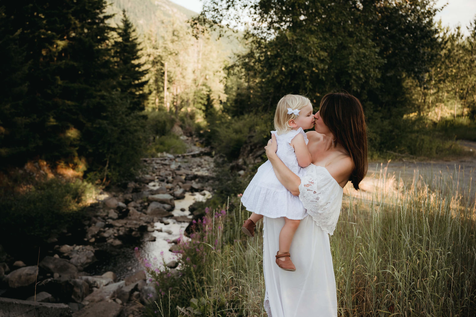 Seattle summer maternity photoshoot: expectant mom in flowing white off-shoulder dress kisses toddler daughter in matching white dress beside a forest creek and Cascade foothills—Lana Sky Photography.