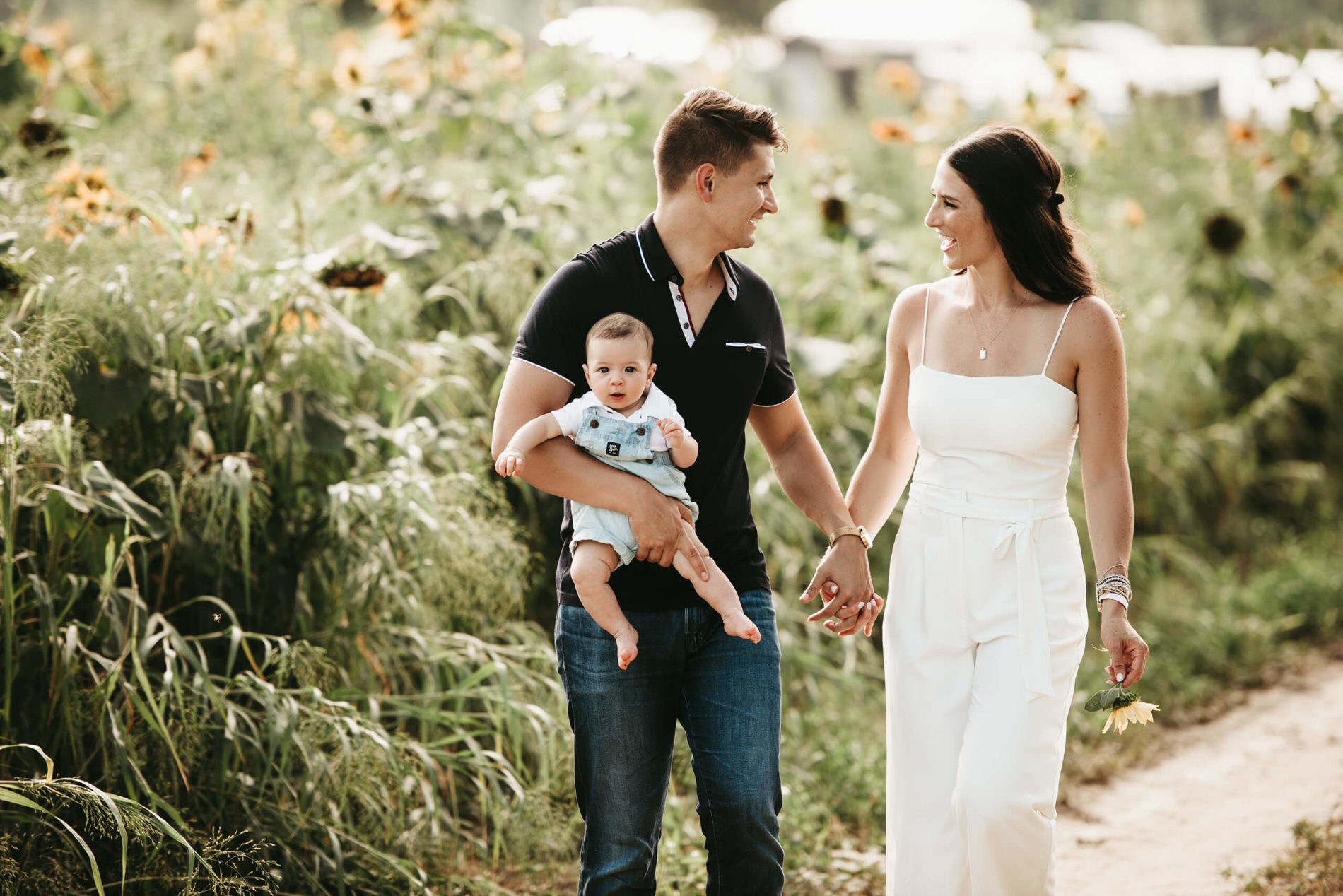 Happy husband and wife holding hands walking through sunflower field with their toddler