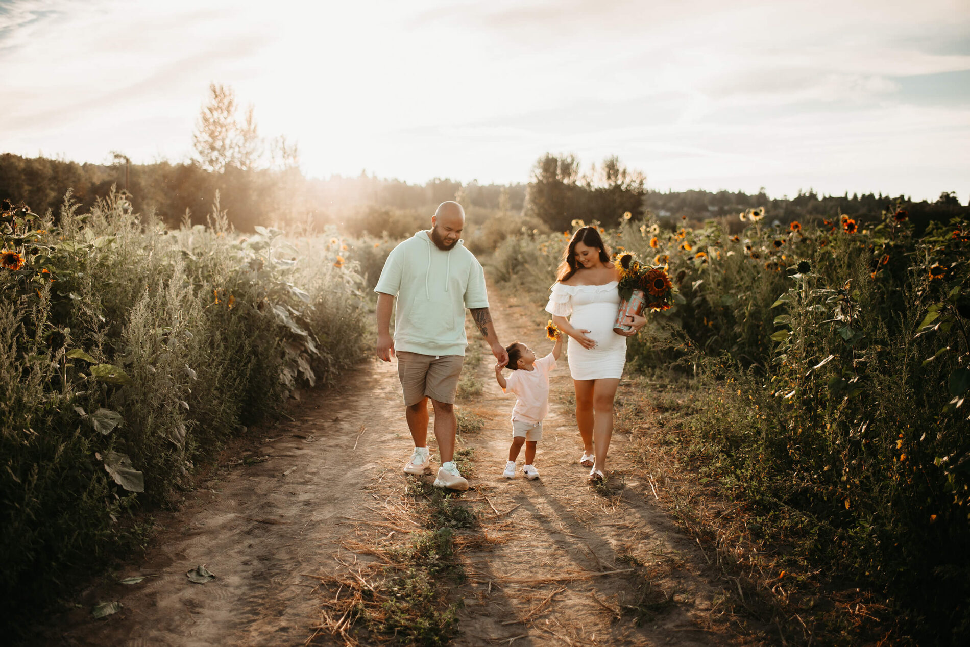 Husband and pregnant wife walking through a sunflower farm with their toddler son