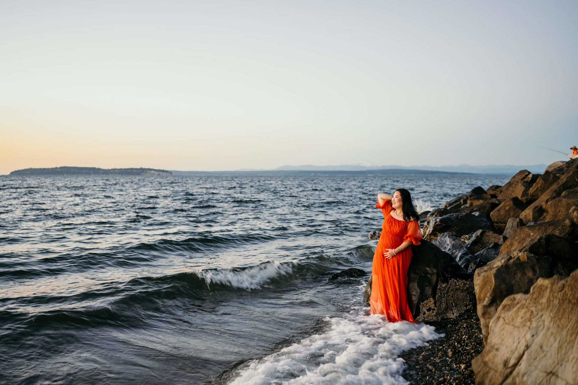 Pregnant woman in a red gown standing on a beach during sunset with waves crushing all around her