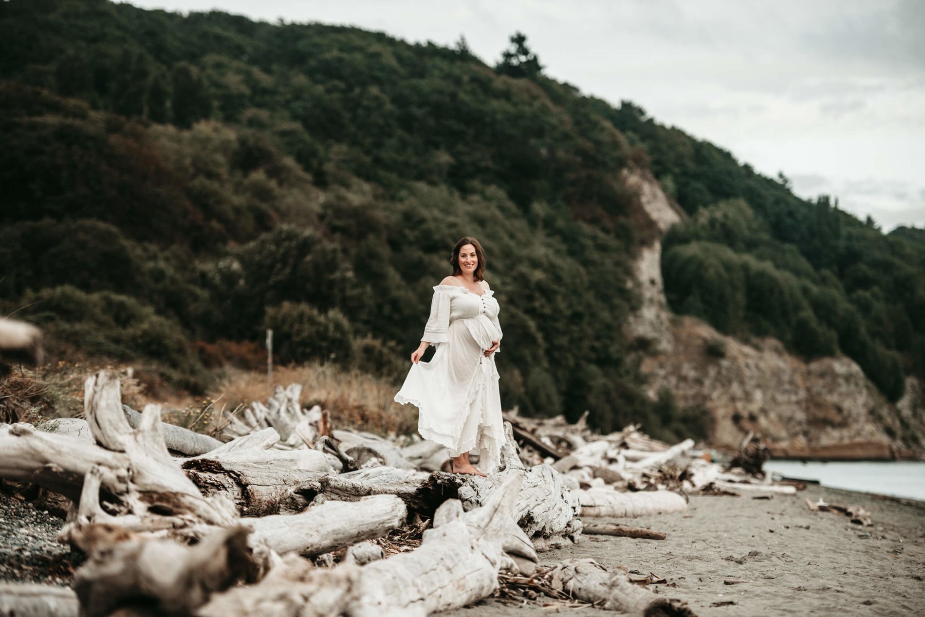 A pregnant woman in a beautiful white dress standing on a fallen tree at a Seattle area beach