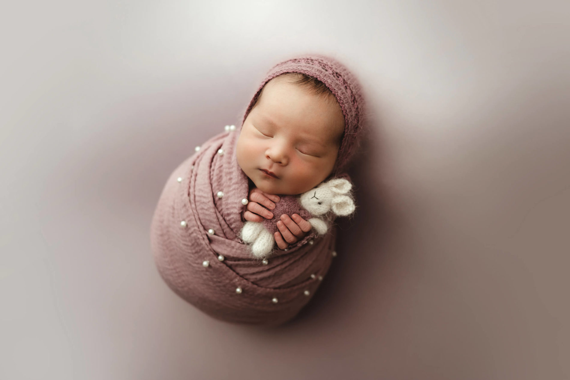 A sleeping newborn girl during photo shoot masterfully wrapped and wearing a headband, holding a stuffed rabbit toy